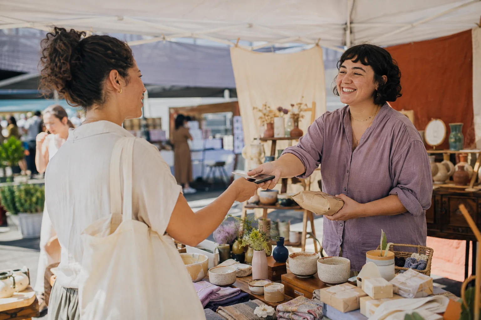 A craft-business owner making a first sale across a colorful neighborhood market table with a smiling customer.